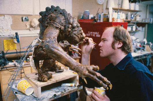 Tippett on the right painting a medium-sized model of the Rancor monster. The monster has a hunched back with bumps, long fingers, and claws, and a chunky head. Its color pallet is black, brown and dirt yellow.