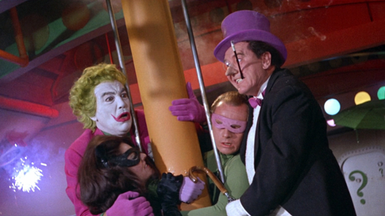 The Batman villains are assembled around a pole in a colorfully-painted building. The Joker, Catwoman, the Riddler, and Two-Face have worried looks on their faces, or appear to have passed out.