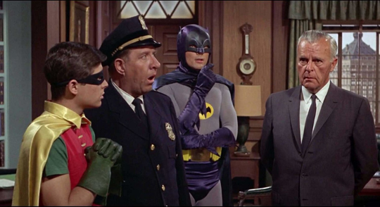 Batman and Robin are meeting with two representatives of the police force in a primarily brown-shaded office room.