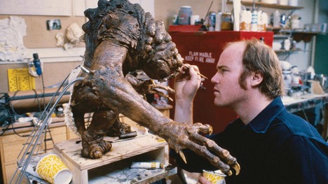 Tippett on the right painting a medium-sized model of the Rancor monster. The monster has a hunched back with bumps, long fingers, and claws, and a chunky head. Its color pallet is black, brown and dirt yellow.
