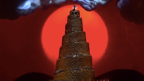 A golden tower with a circular stairway in front of a glowing red sun that marks a blood red sunset. A figure stands on top of the tower. Grey and black clouds are in the sky.