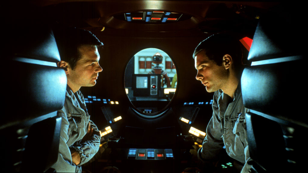 Two men speak in a space capsule while a computer monitor and a spacesuit are visible through a window in the background.