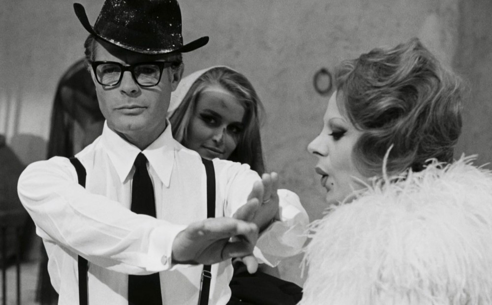 A black-and-white image of a man wearing a black hat and eyeglasses holding his fingers out to a woman in a frilly white dress for her to blow on them.