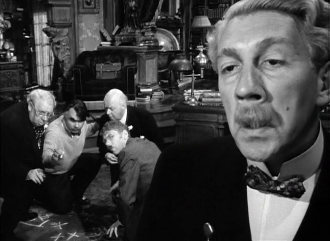A black-and-white image of a mustached man (Richard Haydn) in a suit and bowtie standing in the foreground, while four other men watch him and make chalk markings on the ground in the background.