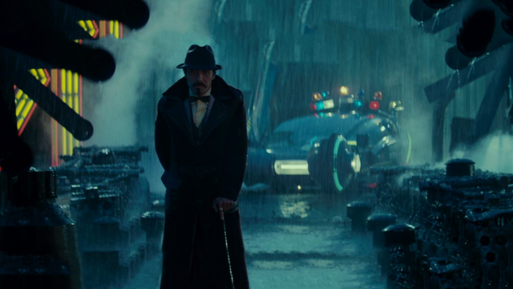A man in a black overcoat and fedora, carrying a cane, stands in an alley during a rainstorm, with a futuristic car visible behind him. 