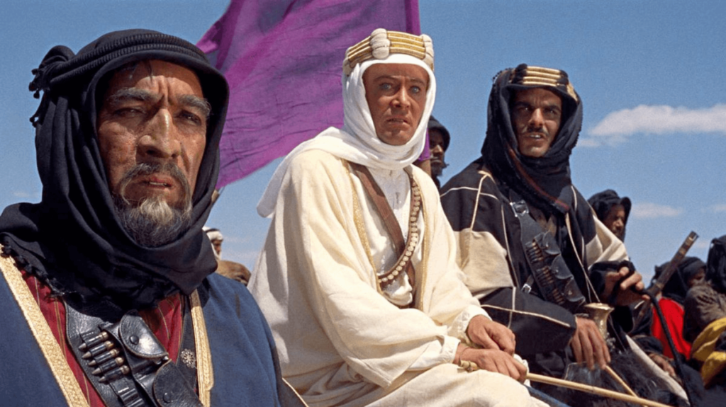 Three men in blue, white, and black robes sit on horseback staring at something offscreen, a purple flag waving behind them. 