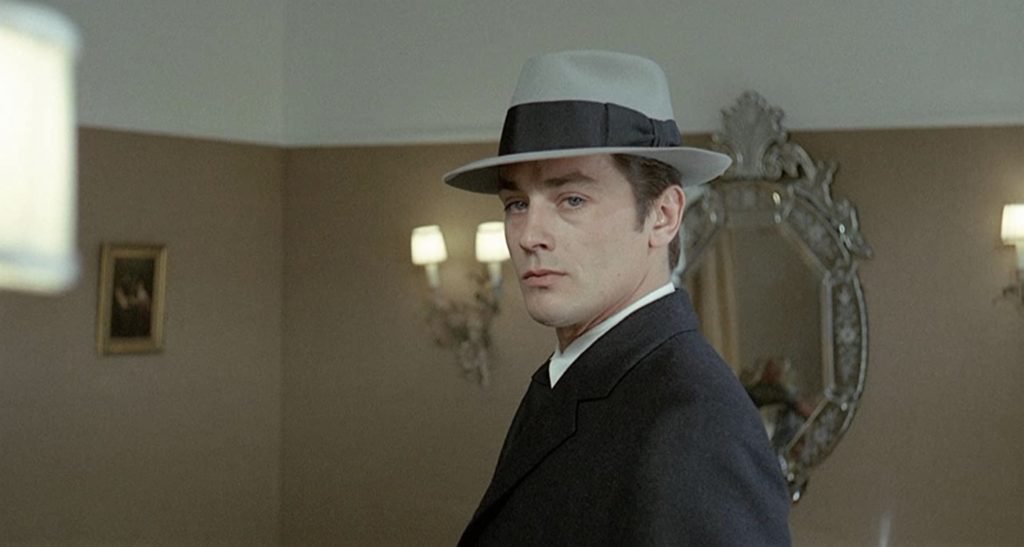 A man in a gray fedora looks near the camera in an elegant room with wall sconces and a mirror behind him.