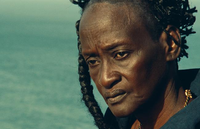 A Black woman looks offscreen at something, the sea stretching out in the background.