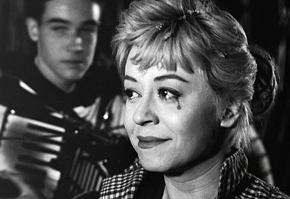 A black-and-white image of a woman shedding a tear as a man plays the accordion behind her.