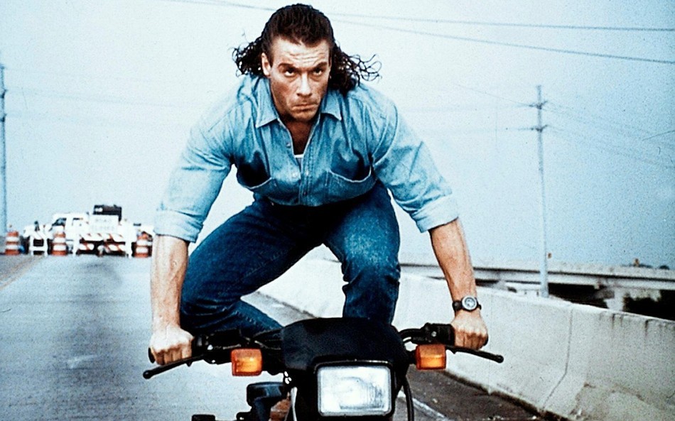 A man in a blue denim shirt crouches on the seat of a moving motorcycle.