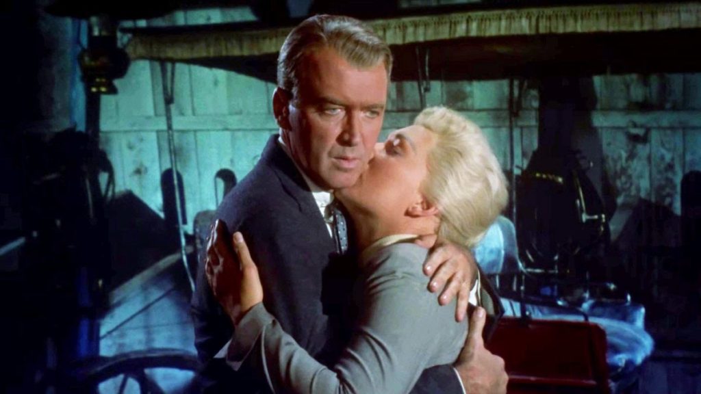 A man (James Stewart) and a woman (Kim Novak) hold each other close in an urban setting with greenish light on the walls behind them.