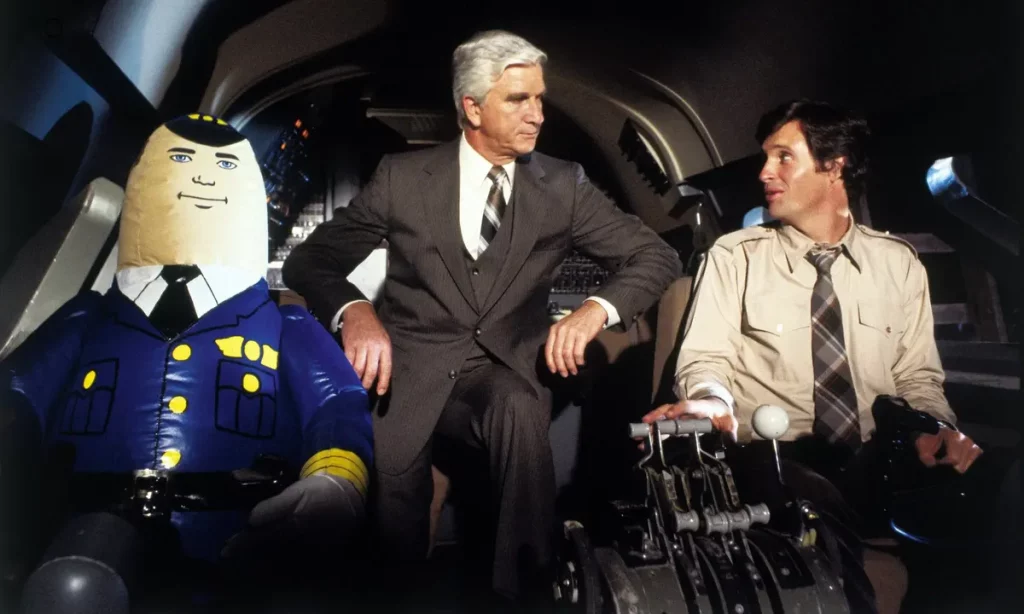 In an airplane cockpit, an inflatable doll is positioned next to a man standing in a suit and the pilot.