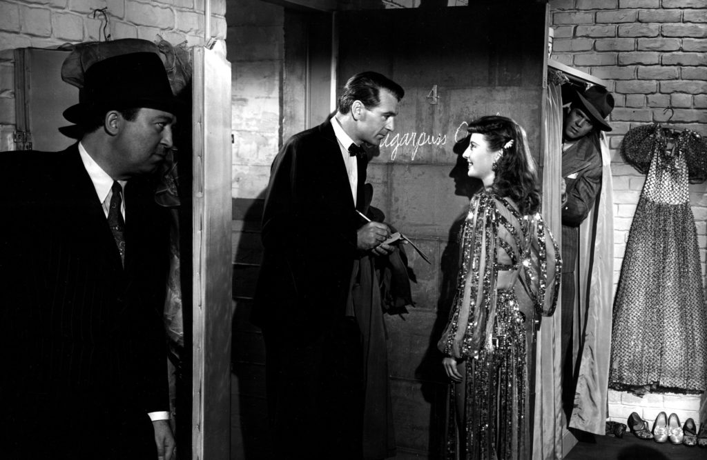 A black-and-white image of a man in a tuxedo (Gary Cooper) and a woman in a sequined dress (Barbara Stanwyck) conversing at an open doorway while two other men eaveswdrop while hiding. 