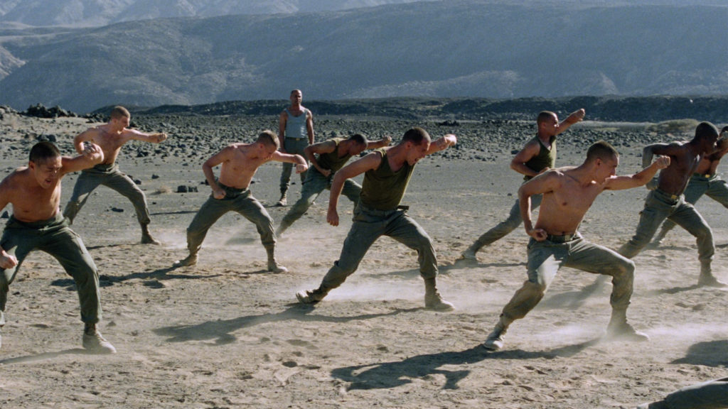 A group of men, some of them shirtless, move synchronously in the desert as another man watches them.