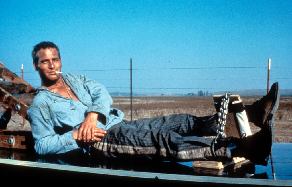 A man in a blue denim shirt wearing ankle chains lies on a black surface, smoking a cigarette.
