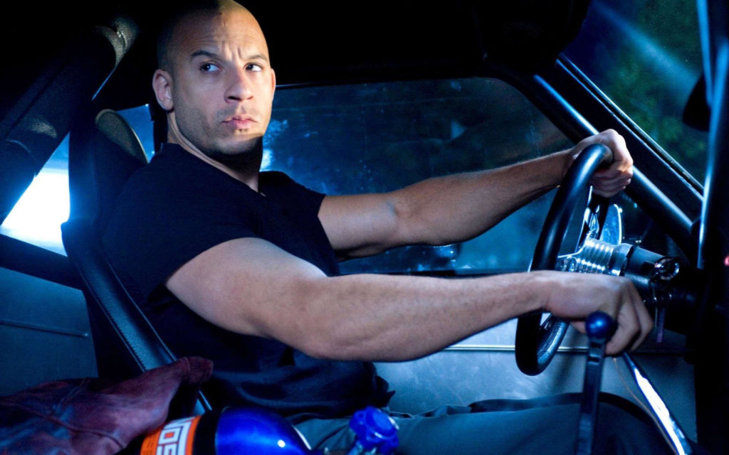 Screengrab from the 2009 film Fast & Furious. Vin Diesel, a bald muscular man dressed in a black t-shirt and blue jeans, is sitting in the driver’s seat of a car, holding the steering wheel and gear shift with each of his hands and looking out the passenger window.