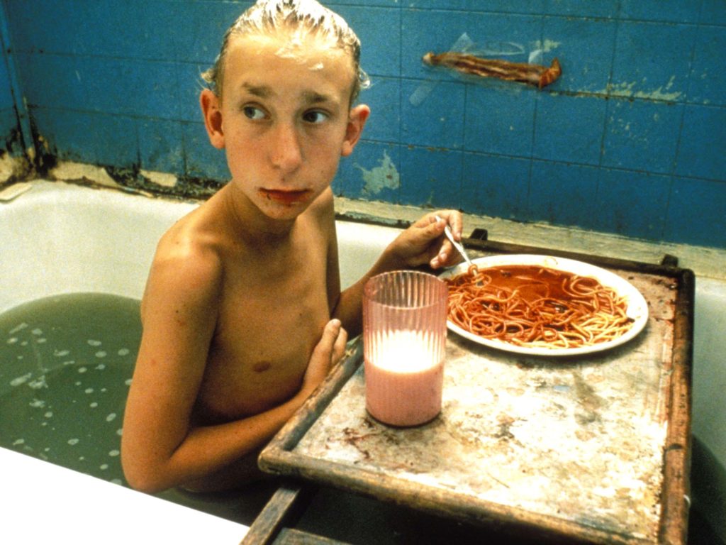 A skinny young boy in a dingy bathtub with blue tiles and dirty water eats spaghetti, a glass of milk on a tray in front of him.