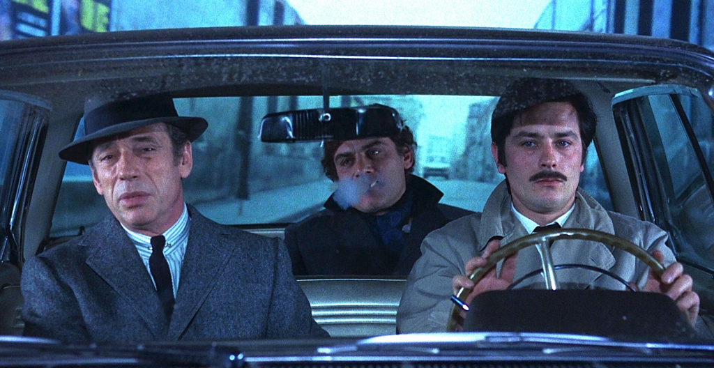 Three men are seated in a car, the man in the backseat smoking a cigarette.