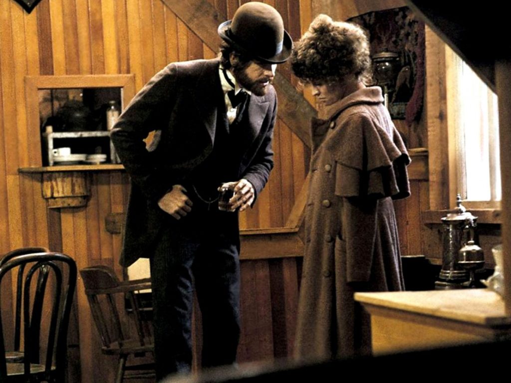 A brown-tinted image of a man in a black suit talking to a woman in a brown coat in a small room with wooden walls.