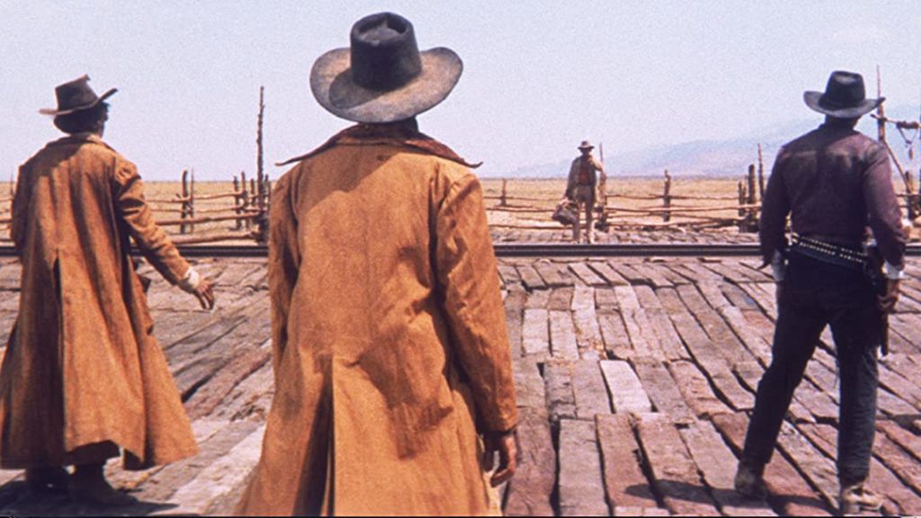 In a remote desert setting, three men wearing cowboy hats and duster jackets with their backs to the camera stand with their hands poised over their guns; a fourth man stands in the distance. 