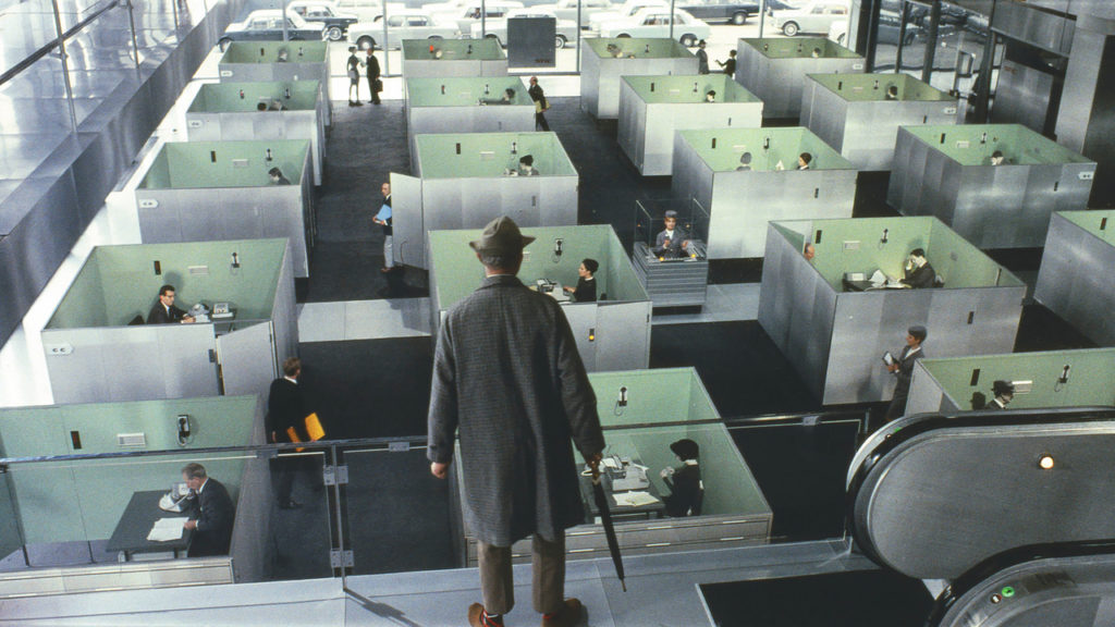 A man in a suit and hat holding a closed umbrella looks down at a network of office cubicles from a higher level.