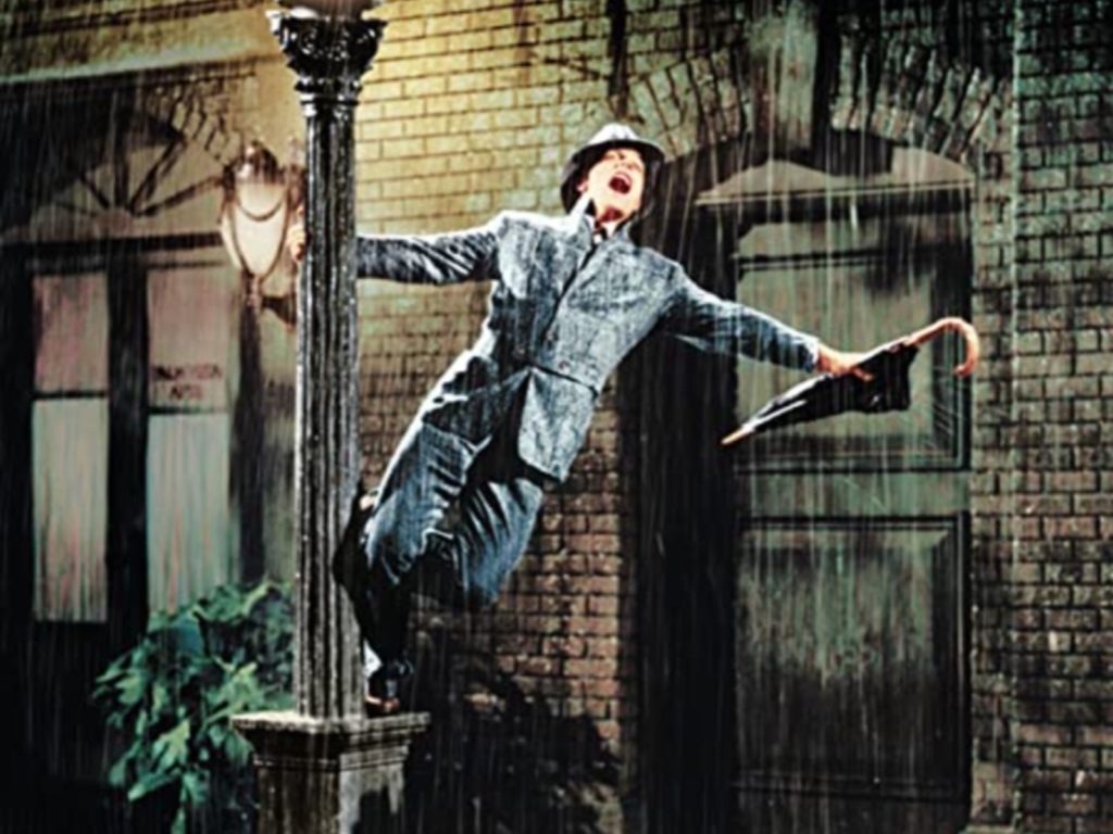 A man stands on the base of a streetlight in a rainstorm, happily singing and holding a closed umbrella.