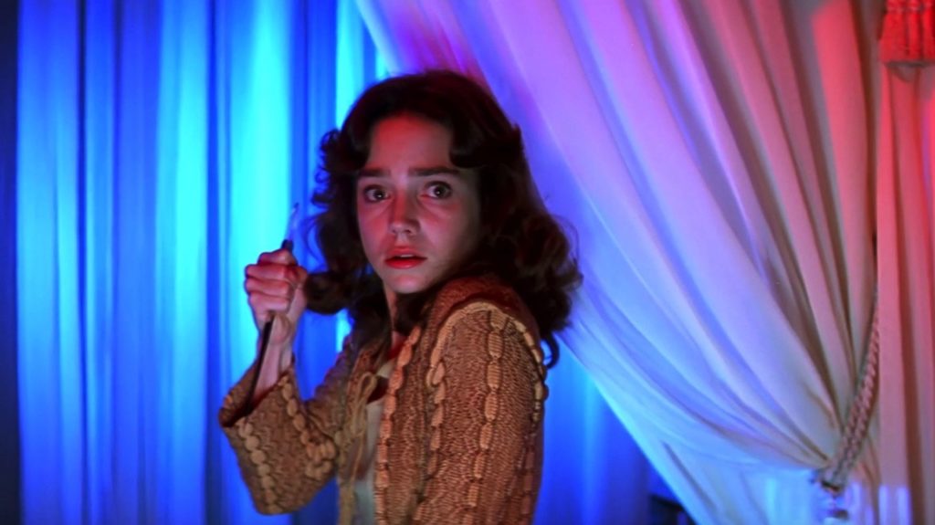 A fearful woman holds a knife as she stands in front of a curtain with blue, purple, and red light cast behind her.