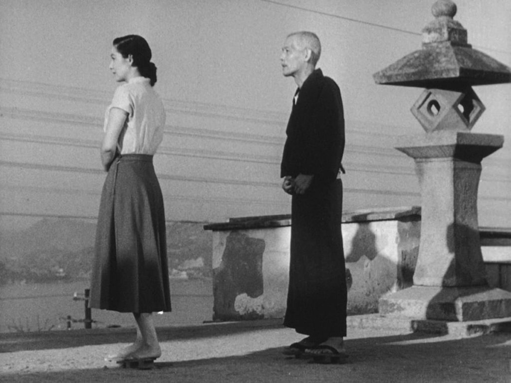 A black-and-white image of a young woman and an old man standing side-by-side, a body of water in the distance.