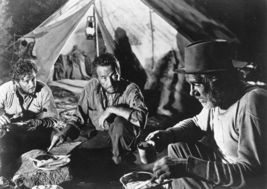 A black-and-white photo of three men sitting around a campfire at night, with a tent pitched behind them. 
