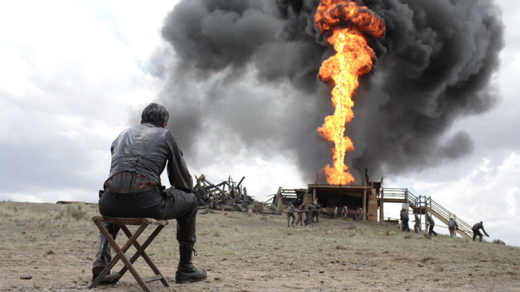 A man seated on a chair, his back to the camera, watches a stream of fire and smoke emerge from an oil well. 