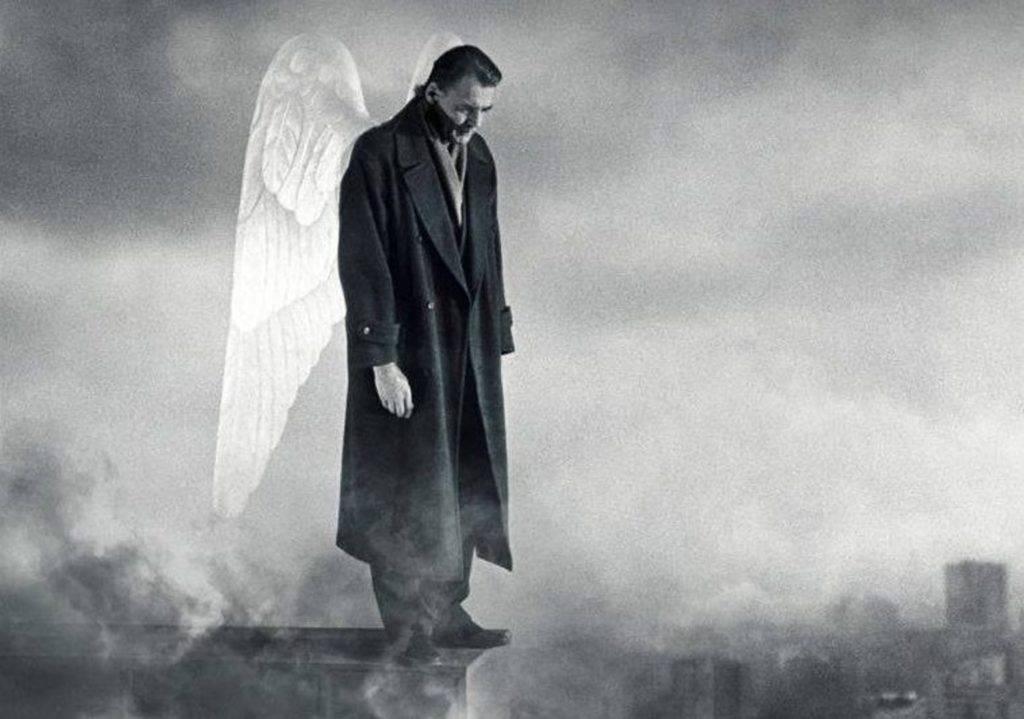 A black-and-white image of a man in an overcoat with angel wings standing on a ledge, the cloudy sky behind him.