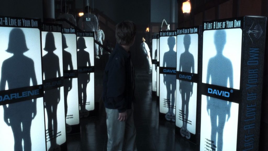 David is center frame, with his neck turned looking backward. To his left are boxes with the shadowy outline of a girl robot and the name, in light-blue text, “Darlene.” To David’s right is the shadowy outline of a boy robot and the name “David.” At the top of both boxes is the text: “AT LAST—A LOVE OF YOUR OWN.”