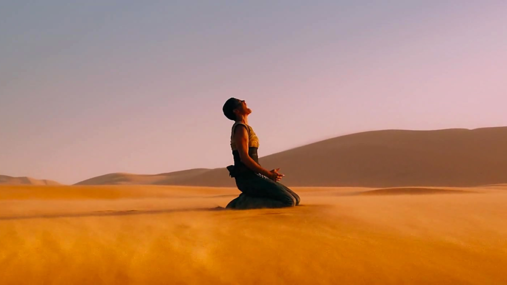 Furiosa (Charlize Theron) kneels in the desert sand, crying. A large mountain looms in the background.