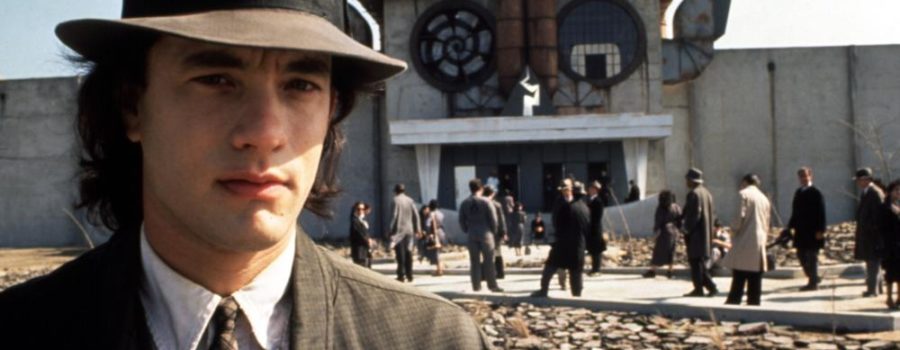 Tom Hanks stands, wearing a hat, in front of an odd building with people loitering in front of it in the film "Joe Versus the Volcano."