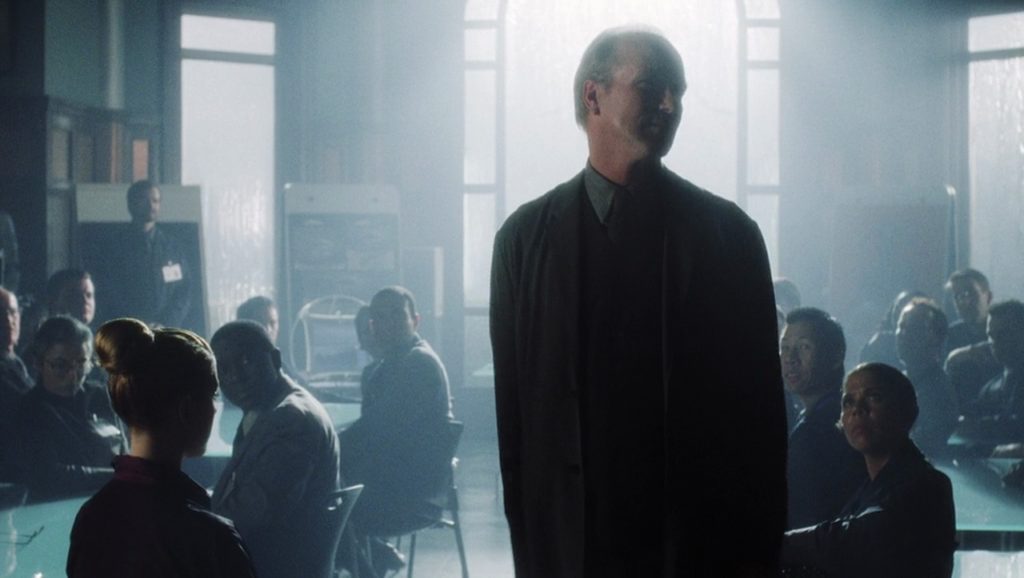 Professor Hobby, portrayed by William Hurt, stands in center frame. To his left and right his colleagues at Cybertronics look up at him with admiration and even awe. Light streams through a large window in the background, and the shot’s cinematography makes ample use of bloom.
