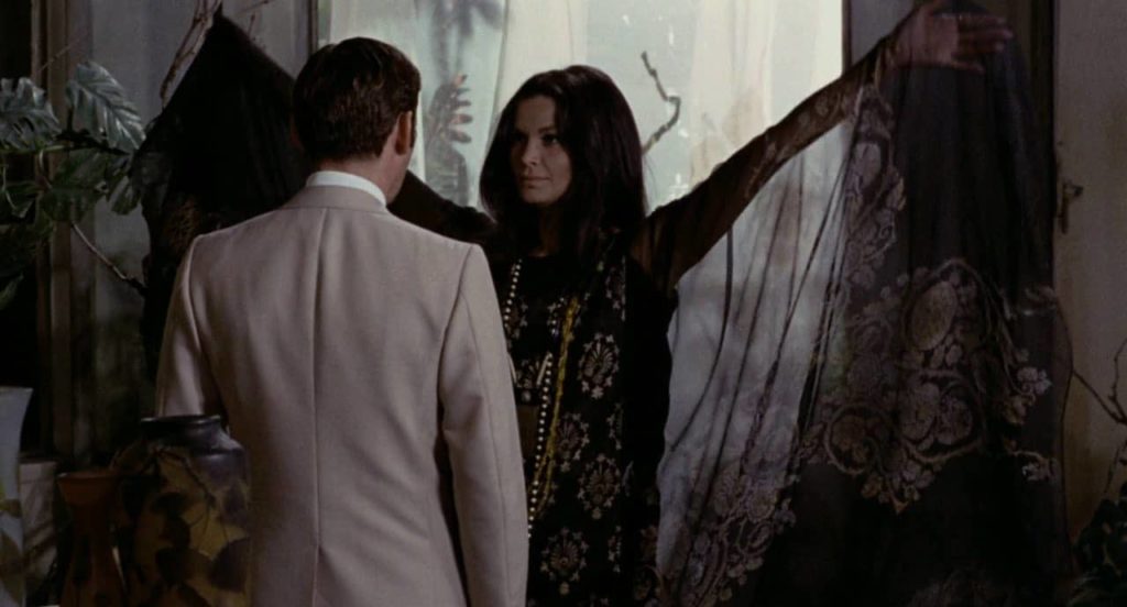 In a handsomely decorated room, a woman raises the arms of her black dress while standing before a man in a beige suit.