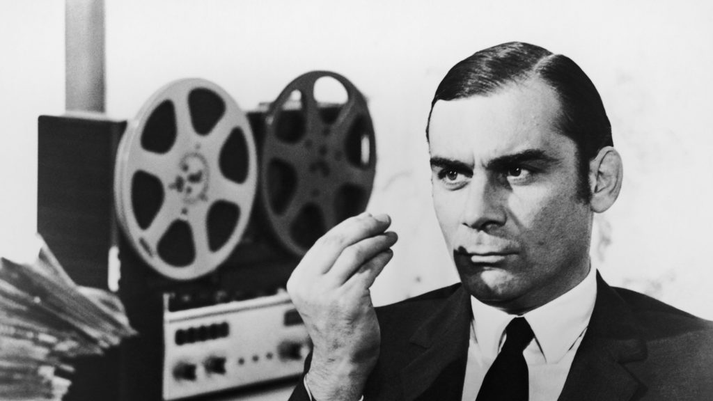 A black-and-white still from "Investigation of a Citizen Above Suspicion" in which a man in a suit looks pensively next to a reel-to-reel audio player.