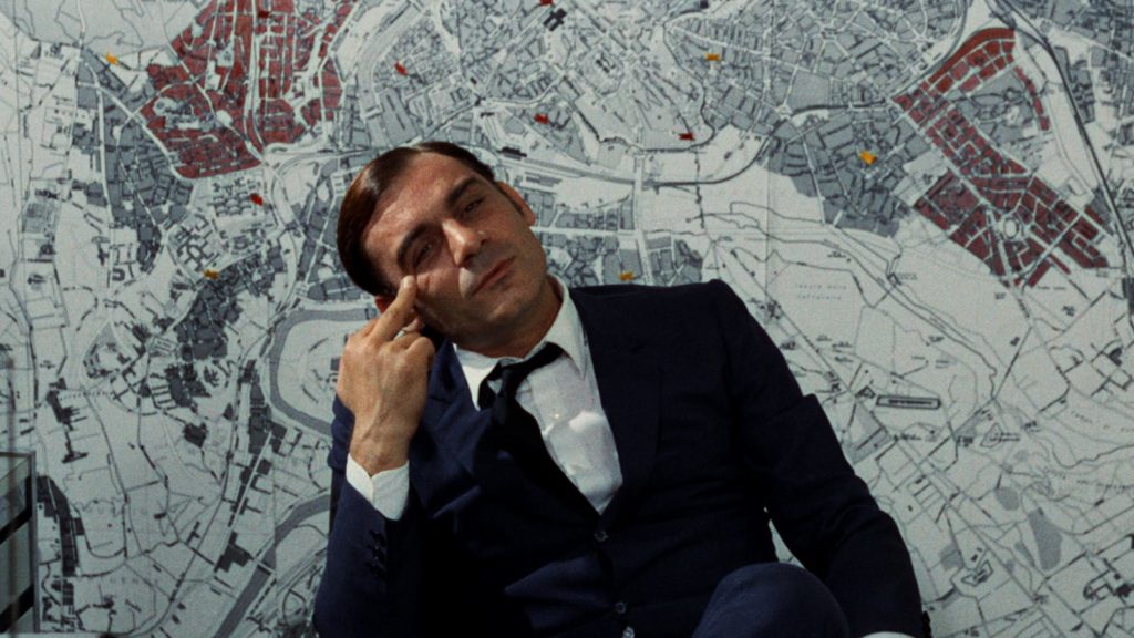 A man in a blue suit sits in front of a wall-sized map with a bored expression on his face. 