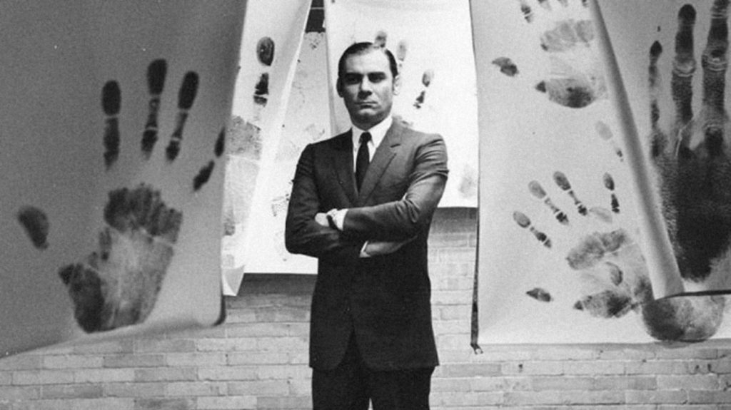 A black-and-white photo of a man in a suit, arms crossed, standing in a room in which large handprints can be seen on sheets hanging from the ceiling. 