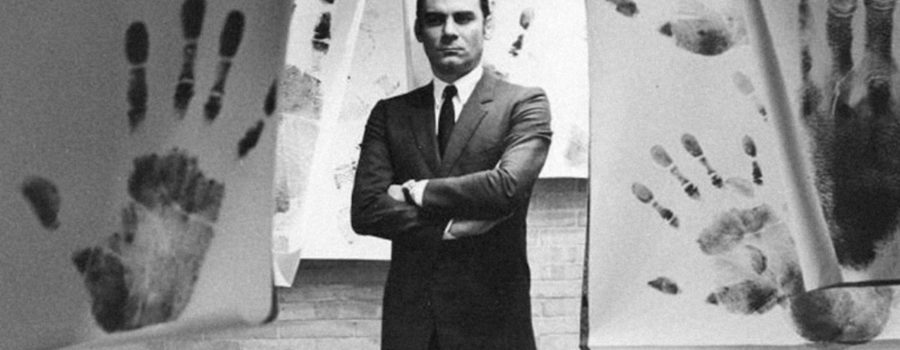 A black-and-white photo of a man in a suit, arms crossed, standing in a room in which large handprints can be seen on sheets hanging from the ceiling.