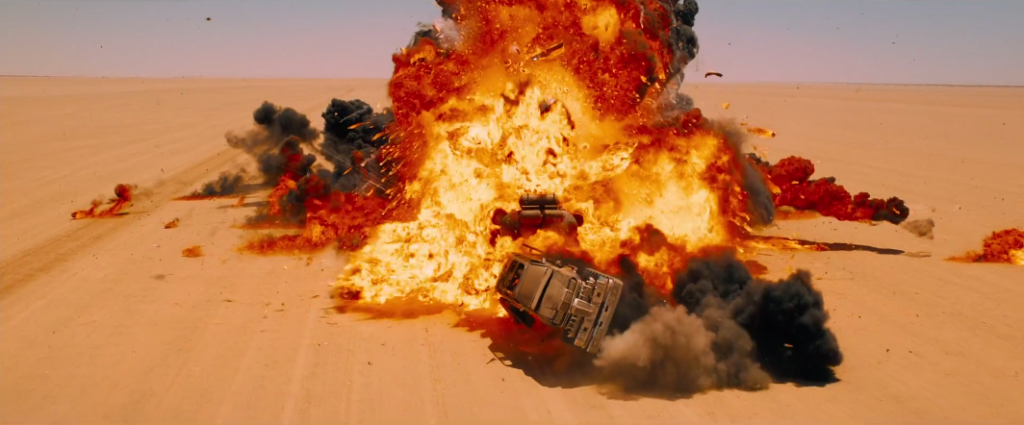 A large, fiery explosion erupts in the center of the desert. There is a large vehicle inside it, and a car flying toward the viewer. Behind it, smoke billows toward the sky.