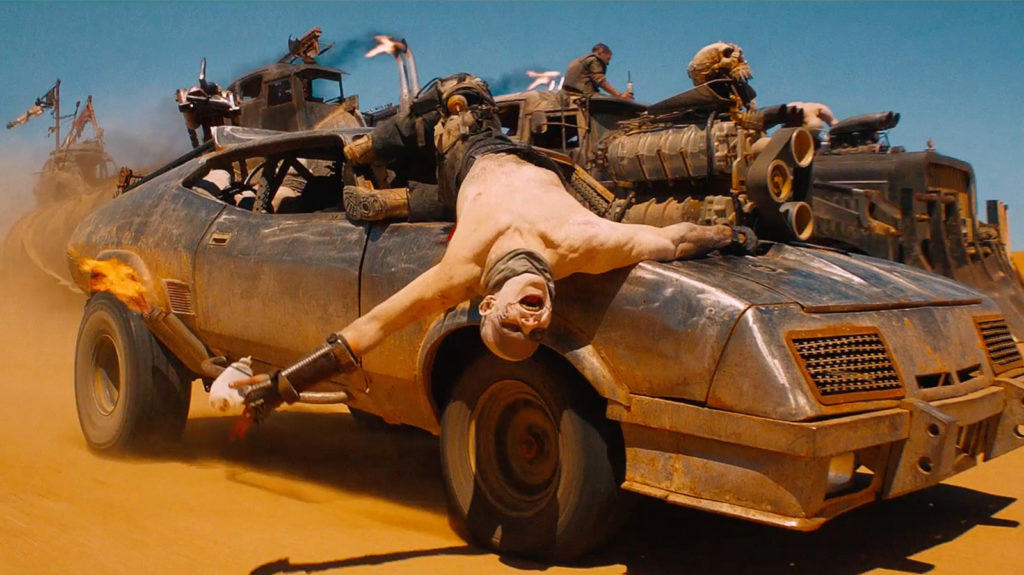A shirtless man lies on his back on the hood of a moving car, holding something flaming in his left hand. Other vehicles are visible in the background, moving across the desert. 