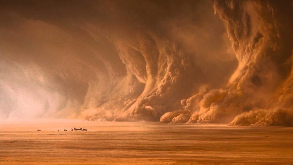A large, wave-like sandstorm approaches the desert from the right. Several tiny vehicles approach it.