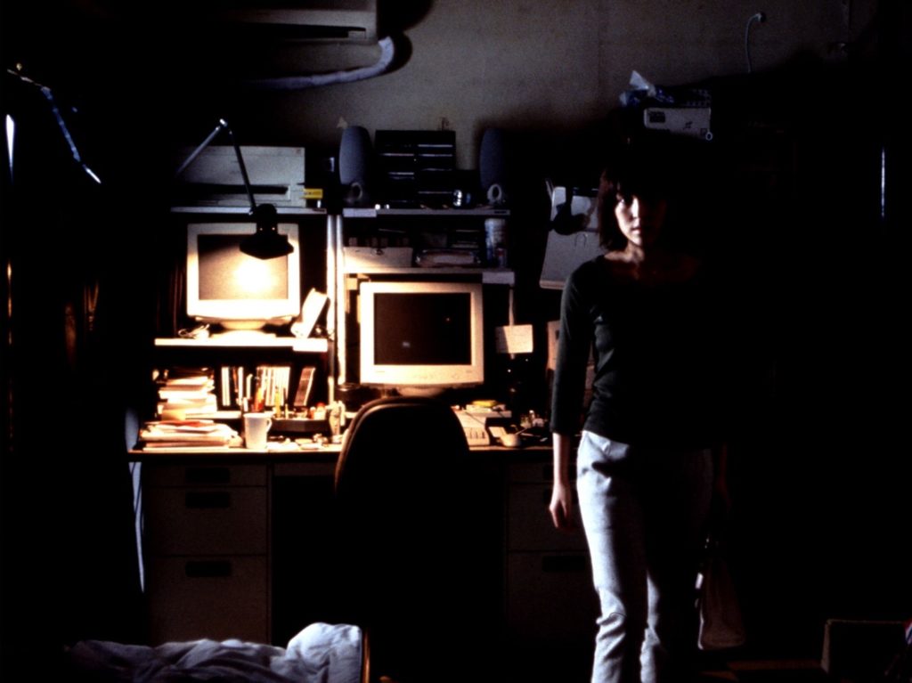 A woman stands in front of a desk with two computer monitors and various office supplies as a lamp casts dim lighting.