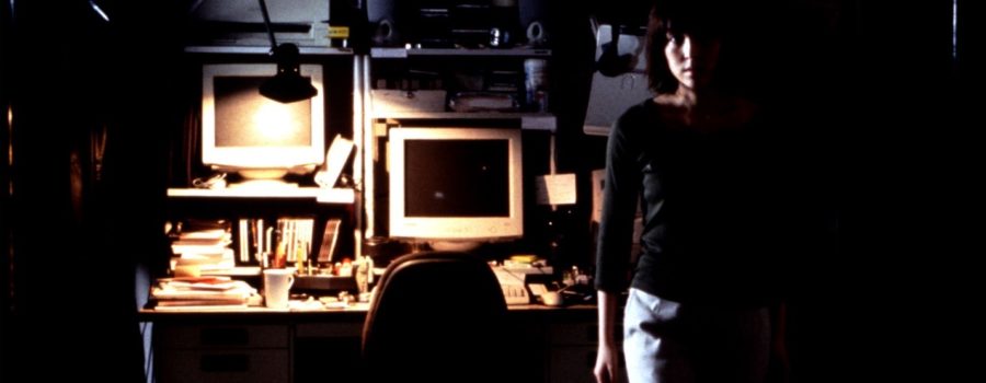 A woman stands in front of a desk with two computer monitors and various office supplies as a lamp casts dim lighting.