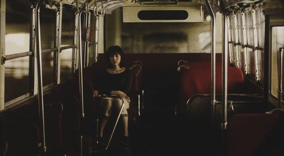 A woman sits on a bus as a street can be viewed passing by through the windows. 