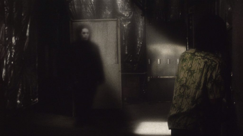 A man in a Hawaiian shirt cowers to the right as a ghost with a pale face approaches him in a shadowy room with a single beam of sunlight.