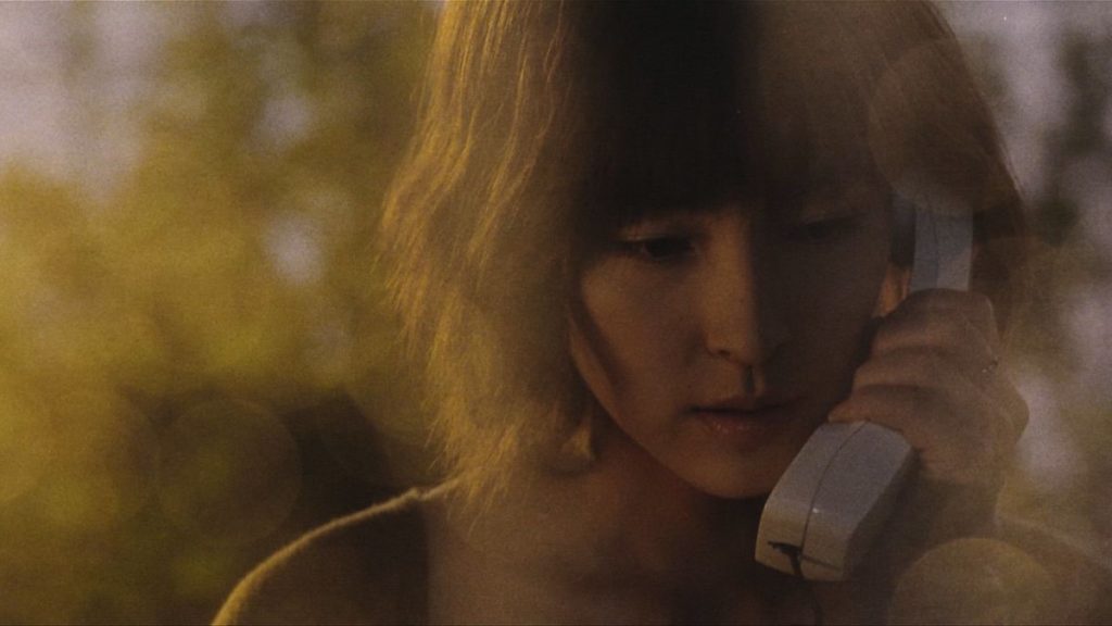 A woman speaks on a telephone as plants and sunlight can be seen behind her.