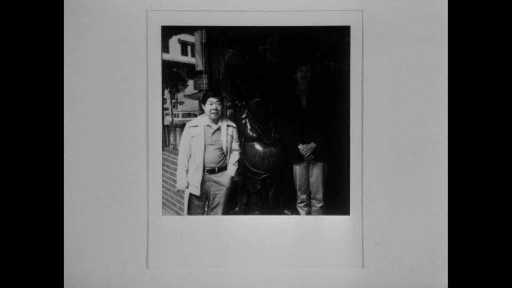 A Polaroid photo of Jo and Chan standing together with a statue of Buddha. A shadow from a nearby awning is cast over the left side of the photo, obscuring Chan’s face and some of the statue.