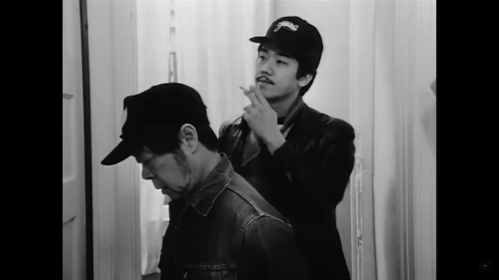 Jo and Steve stand outside of a door in an apartment hallway. Jo looks down and Steve is seen smoking a cigarette.